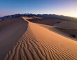© Arek - Expansive desert scenery featuring sun-kissed sand dunes, rolling towards a mountain range under a twilight sky. Peaceful and calm vista