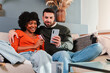 © Jose Calsina - A couple shares a delightful moment while shopping online, using a smartphone and credit card on their cozy couch, showcasing the joy of digital shopping together in the comfort of home.