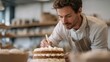 © CYBERPINK - A man wearing a beige apron and working in a bakery. he is holding a wooden spoon and is in the process of decorating a cake. the cake is on a white cake stand and is placed on a table.