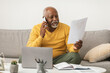 © Prostock-studio - A man in a yellow sweater is seated on a sofa in a cozy living room. He is talking on the phone while examining papers and using his laptop.