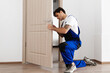 © Prostock-studio - A young male locksmith in a blue uniform is kneeling while carefully installing a door knob on a wooden entrance door. The setting is a clean and modern interior, ideal for professional repairs.