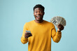 © Prostock-studio - Emotional handsome african american young man in yellow using modern cellphone and showing bunch of cash on blue studio background, recommending newest mobile application, copy space