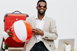 © SHOTPRIME STUDIO - Smiling man with dark skin, sunglasses, beige blazer, white shirt, holding a beach ball, ready for vacation, in front of travel luggage, studio shot, lifestyle concept.