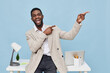 © SHOTPRIME STUDIO - Smiling man with dark skin, wearing a beige sweater, pointing to the side while standing in a bright modern office with a desk, laptop, and decorative plant against a blue background. People lifestyle