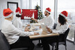 © sofiko14 - Happy team of diverse business people in Santa hats listening their middle aged male bearded chief, showing dynamics of r world market, using digital TV screen at office decorated with Christmas tree.