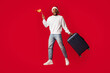 © Prostock-studio - A lively traveler wearing a santa hat jumps joyfully holding a small flag and pulling a suitcase on wheels. The bright red background adds energy to the scene.