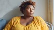 © Maryna - Woman with a large bust is sitting on a couch in a yellow shirt. She has a serious expression on her face