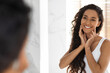 © Prostock-studio - A woman stands in a bright bathroom smiling at her reflection. She has long curly hair and is touching her face, enjoying the moment after applying skincare products.