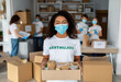 © Prostock-studio - A young female volunteer in a medical mask smiles while holding a box of canned food and grains. She is working at a charity center with diverse colleagues during a community service activity.
