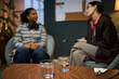 © DragonImages - Middle aged Caucasian woman interviewing young Black woman in office setting, both seated and engaged in conversation, glasses of water and notepad on table in foreground