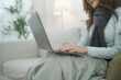 © DG PhotoStock - Cozy work from home lifestyle of a woman using laptop while sitting on sofa in a warm living room during winter, representing remote work, freelance job and comfortable home office environment.