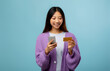 © Prostock-studio - A young woman stands smiling as she holds a smartphone in one hand and a credit card in the other. She appears happy and engaged, set against a light blue backdrop.