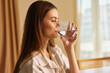 © SHOTPRIME STUDIO - Woman drinking water from a glass at home, portrait side view of a female in morning light. Hydration, healthy routine and relaxation for wellness and daily self care.