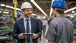 © ChristMasX - A smiling manager in a suit and hard hat discusses something on a tablet with an employee in a factory setting.