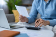 © Liubomir - Man holding a paper document and using a calculator while sitting at a table with a laptop, managing personal finance, budgeting monthly expenses, and reviewing bills from home