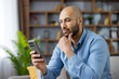 © Liubomir - Young bald bearded man sitting on a sofa at home, focused on his smartphone while thoughtfully browsing and making a serious decision about content or information online