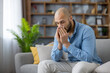 © Liubomir - Young man using a tissue to blow his nose while sitting on a couch at home, experiencing symptoms of a cold, flu, virus, or seasonal allergy and needing relief