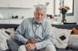 © Daniel - Elderly man sits on a sofa holding his stomach in pain. He struggles with abdominal discomfort and fatigue while trying to relax at home.