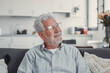 © Daniel - Elderly man sits on a sofa at home looking outside the window. He feels calm and reflective while enjoying a serene moment in his daily life.