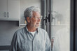 © Daniel - Elderly man stands by the window smiling while looking outside. He enjoys calm, reflective moments and daydreams peacefully about life at home.