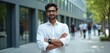 © Vadym - Young Indian man with glasses stands outdoors in front of modern office building. Confident professional smiles arms crossed, wearing white shirt and smart watch. Tech career focus.