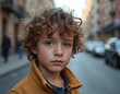 © Pete - Young boy with curly hair looks seriously. He stands on city street with blurred cars and buildings. Child wears yellow jacket and blue shirt. Natural expression, outdoor portrait.