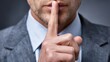 © Formoney - A man dressed in a suit places his index finger against his lips, signaling others to be quiet.