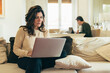 © Lomb - Middle aged woman working from home using laptop in living room with partner in background