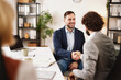 © Stockphotodirectors - Two team members shake hands while sitting at a table. They are surrounded by colleagues who are discussing ideas. The meeting is taking place in a modern office environment.