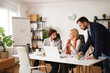 © Stockphotodirectors - Team members are collaborating in an office setting. They are discussing plans, reviewing documents, and sharing ideas around a table with a laptop nearby.