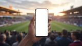 Hand holding smartphone with blank screen at a crowded outdoor sports stadium during sunset