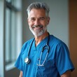 © Viktor - Smiling man doctor grey hair beard blue scrubs stethoscope. Experienced medical professional ready for patient care. Healthcare worker stands in hospital room.