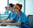 © Viktor - Two young female nurses in blue scrubs work on computer together. They sit at desk typing on keyboard in modern hospital office. Women have dark hair tied up and wear medical uniforms.