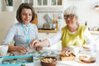 © shurkin_son - Female therapist in geriatric clinic assisting retired woman in glasses to decide jigsaw game or puzzle to overcome dementia consequences playing together at kitchen table, having fun and tea