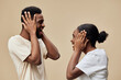 © SHOTPRIME STUDIO - Happy young couple joyfully playing with their hands over their ears, expressing fun and excitement against a soft beige background, ideal for emotional and lifestyle concepts