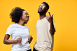 © SHOTPRIME STUDIO - Two joyful young models with afro hairstyles wearing casual outfits and sunglasses, dancing against a vibrant yellow background, expressing happiness and youthfulness