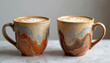 © Maryna - Two unique marbled coffee cups with latte art sit on a counter. The handcrafted mugs feature earthy tones and artistic patterns. They are perfect for a cozy cafe setting or home decor.