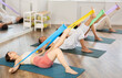 © JackF - Concentrated girl and group of sporty people lying on mats and performing set of pilates exercises with resistance bands in modern fitness studio