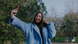 © tol_u4f - Woman takes selfie in park while wearing headphones during daytime in a green outdoor setting surrounded by trees