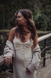 © Sofia - Young woman in a dress and cardigan stands on a wooden bridge by a pond, wind moving her hair, soft daylight, muted green tones, spontaneous mood