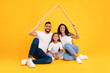 © Prostock-studio - A joyful family sits together on the floor, with a young girl in the middle, holding a cardboard roof above their heads. They wear matching white shirts and jeans, smiling widely.