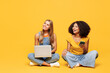 © ViDi Studio - Full body two young friends buddies IT women wear denim casual clothes sit together hold work on laptop pc computer use mobile cell phone isolated on plain yellow orange background. Lifestyle concept.