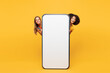 © ViDi Studio - Full body two young friends buddies women wearing denim casual clothes together stand behind big huge blank screen area mobile cell phone isolated on plain yellow orange background. Lifestyle concept.