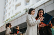 © qunica.com - Two women stand in a modern urban setting, examining a blue folder and discussing plans with colleagues. Nearby coworkers work on laptops, highlighting collaboration in a busy city environment.