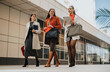 © qunica.com - Three women in chic coats stroll past a modern office facade, chatting and carrying tablets, folders, and bags, showcasing teamwork, style, and urban work life.