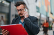 © qunica.com - A man wearing glasses and a blazer speaks on a smartphone while holding a red folder in a colorful urban street, among people. He looks focused as he handles work tasks, a sign of busy associates.