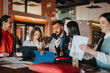 © qunica.com - A diverse group of people sits around a table, discussing documents and sipping drinks in a lively cafe. They share ideas, review charts, and collaborate like a team of coworkers.