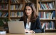 © Ava - Female professor sits in the university library with a laptop, preparing for a lecture. High quality