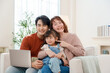 © Ar_TH - Happy Asian family of three, a father, mother, and young daughter, smiling and sitting on a sofa with a laptop at home, enjoying a moment of family bonding and connection.