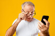 © Prostock-studio - A man wearing glasses is surprised while looking at his smartphone. His facial expression shows shock or amazement. He has short gray hair and is dressed in a white shirt.